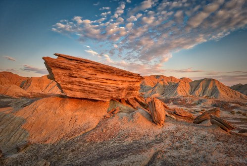A dramatic rock formation bathed in warm sunset light, surrounded by layered hills and a colorful sky.