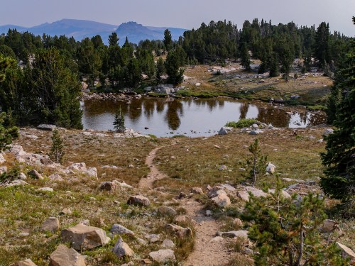 A serene landscape featuring a pond surrounded by trees and rocky terrain, with distant mountains in the background.