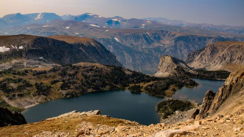 A panoramic view of rugged mountains and two serene lakes surrounded by lush greenery under a clear sky.