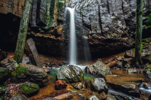 A serene waterfall cascades over rocky cliffs into a tranquil pool, surrounded by lush greenery and large stones.