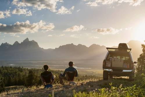 Two people sit on a hillside, overlooking a valley and mountains at sunset, with a parked vehicle nearby.