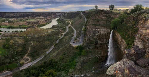 A scenic view of a waterfall cascading down a cliff, surrounded by lush greenery and winding roads along a river.