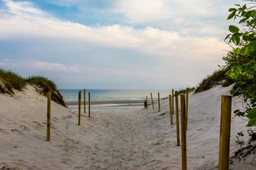 A sandy path between grassy dunes leads to a calm ocean under a cloudy sky.