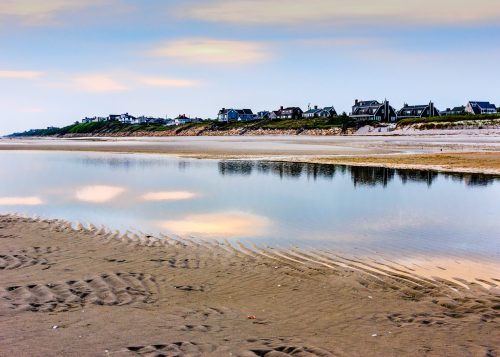 A calm beach scene with a reflective waterline and houses in the background under a soft, pastel sky.