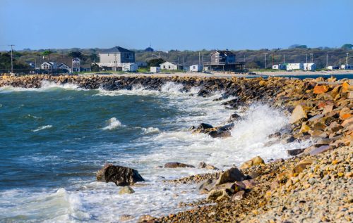 Waves crash against a rocky shoreline with houses in the background under a clear blue sky.