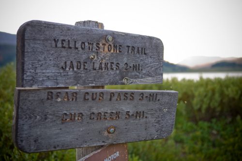 Wooden trail sign with directions to Yellowstone Trail, Jade Lakes, Bear Cub Pass, and Cub Green, near a lake.