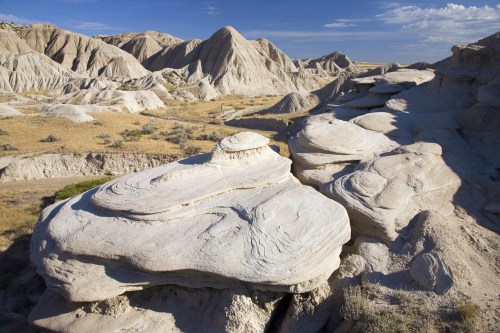 A vast landscape of unique, layered rock formations under a clear blue sky, with rolling hills in the background.