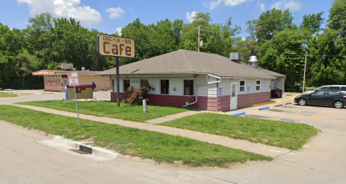 A small café building with a sign, surrounded by greenery and a parking lot, on a sunny day.