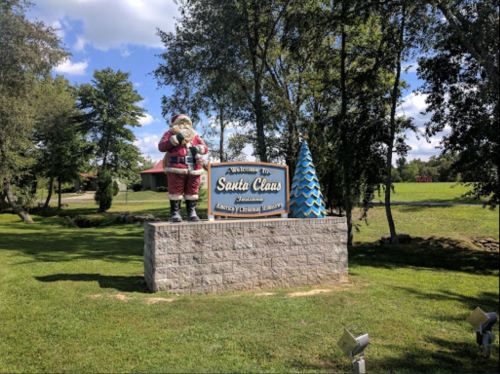 Welcome sign for Santa Claus, featuring a statue of Santa and a blue tree, surrounded by greenery and a blue sky.