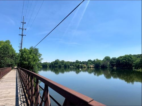 A wooden bridge over a calm river, surrounded by lush greenery and a clear blue sky. Power lines run overhead.