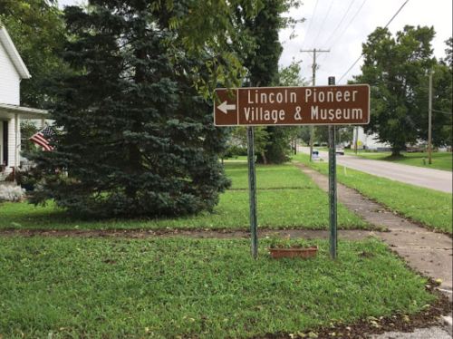 Sign directing to Lincoln Pioneer Village & Museum, with greenery and a residential area in the background.