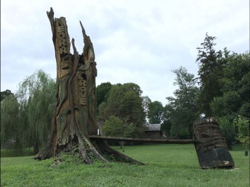 A carved tree stump with the words "Cedar Grove" and a large wooden sculpture in a grassy area.