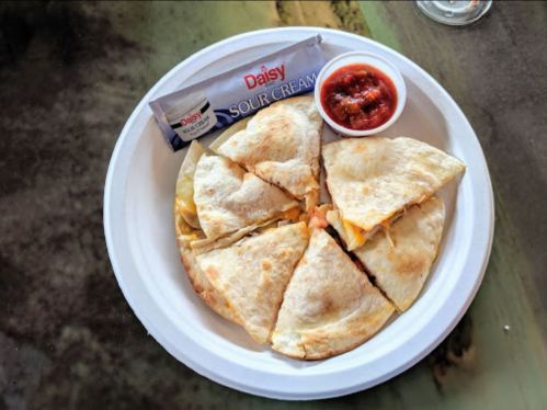 A plate with sliced quesadilla, a cup of salsa, and a packet of sour cream on a table.
