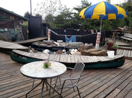 A wooden deck with a table and chair, a canoe, rocks, and a large yellow and blue umbrella in a rustic outdoor setting.