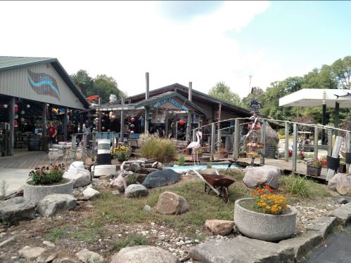 A rustic shop with a wooden bridge, surrounded by rocks and flowers, set in a scenic outdoor area.