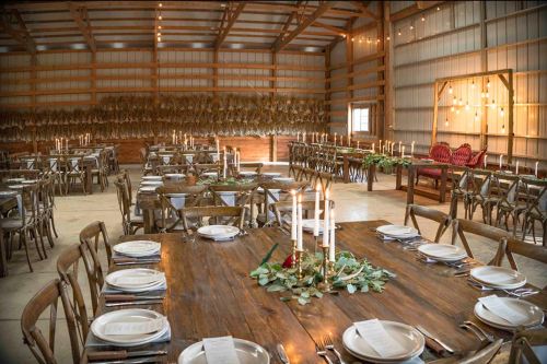 A rustic barn interior set for a dinner, featuring wooden tables, candles, and greenery decor.