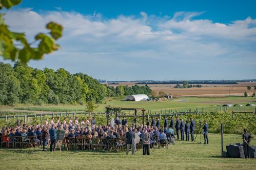 A scenic outdoor wedding ceremony with guests seated on grass, surrounded by trees and fields under a blue sky.