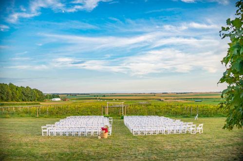 Outdoor wedding setup with white chairs arranged in rows, surrounded by green fields and a blue sky.