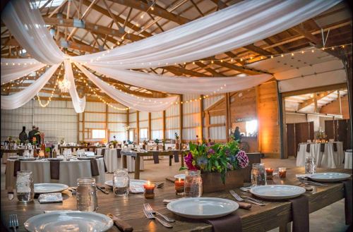 A beautifully decorated barn interior with tables set for dining, draped fabric, and soft lighting creating an elegant atmosphere.