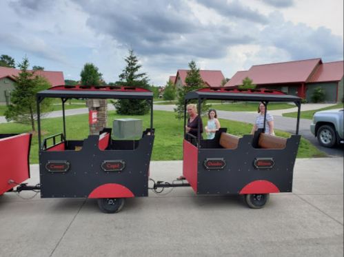 A small train with two cars parked on a driveway, with three people standing beside it and trees in the background.
