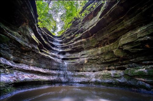 A serene waterfall cascading down layered rock formations into a calm pool, surrounded by lush greenery.
