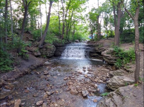 A serene waterfall cascades over rocks, surrounded by lush green trees and a peaceful stream.
