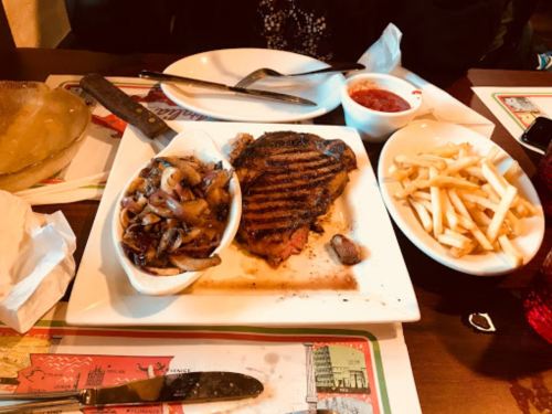 A plate with grilled steak, sautéed mushrooms, French fries, and a small bowl of salsa on a restaurant table.