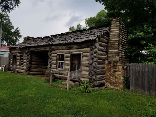 A rustic log cabin with a slanted roof, surrounded by greenery and a wooden fence.
