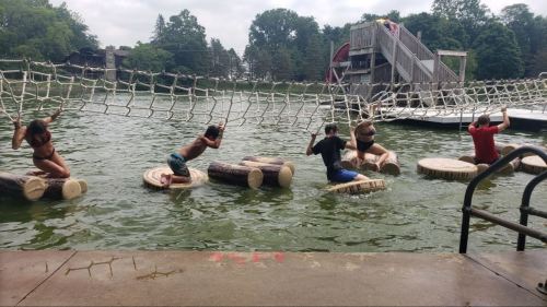 Five people balance on floating logs in a lake, navigating under a rope net in a fun outdoor activity.
