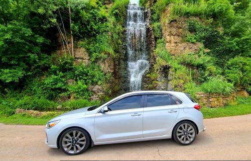 A silver hatchback parked beside a lush green hillside with a cascading waterfall in the background.