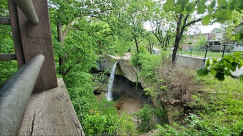A serene waterfall cascades into a lush, green forested area, surrounded by trees and rocky terrain.