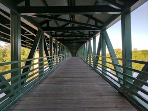 A green metal bridge with a wooden walkway, surrounded by trees and reflecting water on either side.