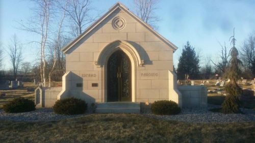 A stone mausoleum with arched entrance, surrounded by grass and trees, in a cemetery setting.