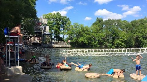 Children playing on floating platforms in a lake, with a rope bridge and a lifeguard watching from the shore.