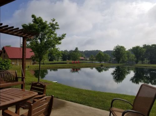 A serene view of a pond surrounded by trees and a grassy area, with a few red chairs in the distance under a cloudy sky.