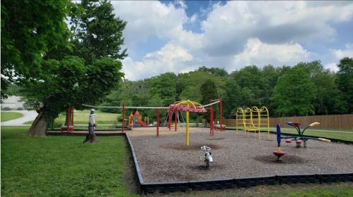 A colorful playground with various equipment, surrounded by green trees and a gravel surface under a partly cloudy sky.