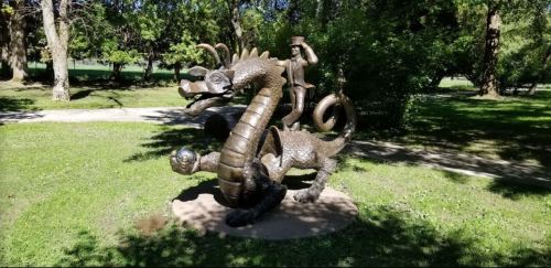 A bronze sculpture of a dragon with a rider, set in a grassy park surrounded by trees.