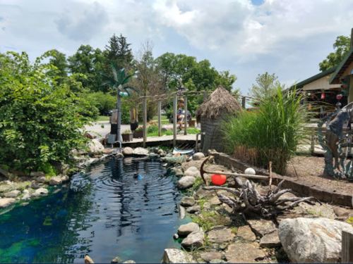 A serene pond surrounded by rocks, greenery, and a thatched hut, under a partly cloudy sky.
