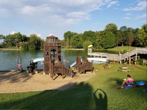A playground by a lake features slides and a dock, with people enjoying the water and relaxing on the grass.