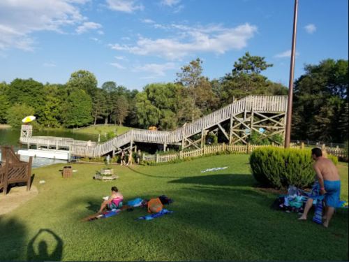 A wooden slide by a lake, with people relaxing on the grass and trees in the background under a blue sky.