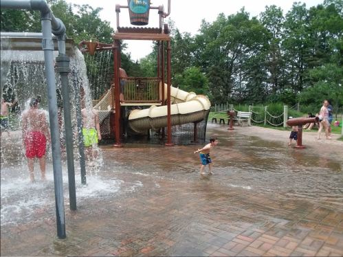 Children play in a splash pad area with water features and a large slide, surrounded by trees and other families.