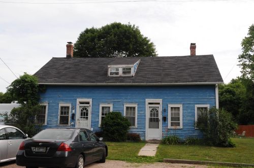 A blue two-story house with a sloped roof, front porch, and a car parked in the driveway. Green shrubs are in front.
