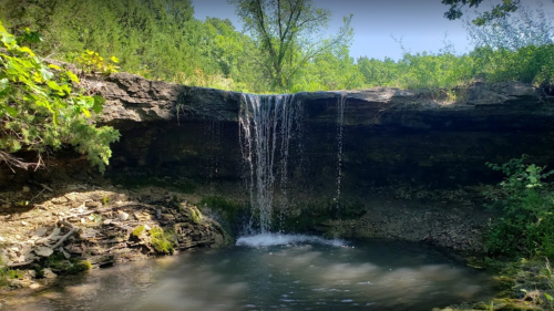 A serene waterfall cascading over rocky cliffs into a calm pool, surrounded by lush greenery and trees.