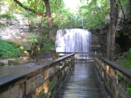 A wooden walkway leads to a waterfall surrounded by lush greenery and rocky cliffs.