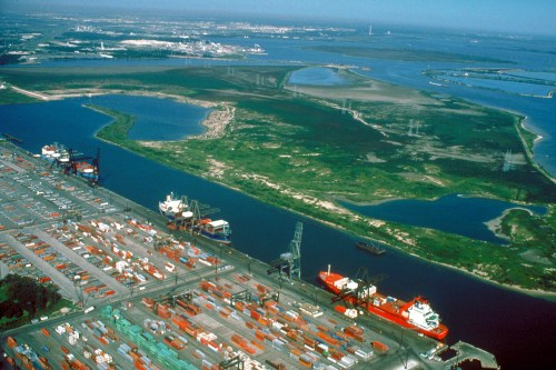 Aerial view of a busy port with shipping containers, cranes, and a river surrounded by green land and distant cityscape.