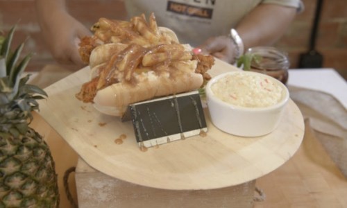 A wooden platter holds two hot dog buns filled with fried chicken and fries, alongside a bowl of coleslaw.