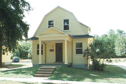 A yellow two-story house with a gabled roof, front steps, and surrounded by greenery on a sunny day.