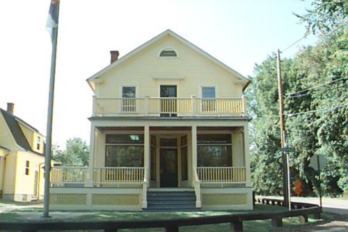 A yellow two-story house with a porch, surrounded by trees and a flagpole in front.