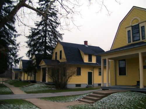 A row of yellow houses with dark roofs, surrounded by trees and a light dusting of snow on the ground.