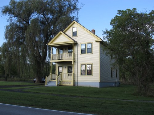 A yellow two-story house with a porch, surrounded by greenery and a large tree, set against a clear blue sky.